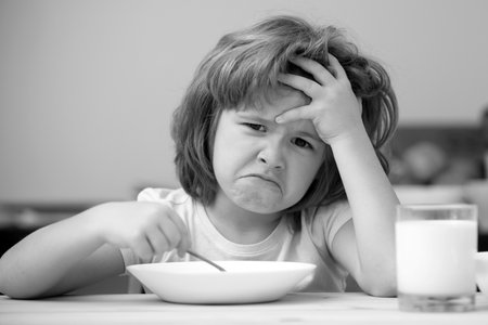 Unfocused little boy having soup for lunch. Unhappy Caucasian child sit at table at home kitchen have no appetite. Upset little kid refuse to eat organic cereals with milk.の写真素材