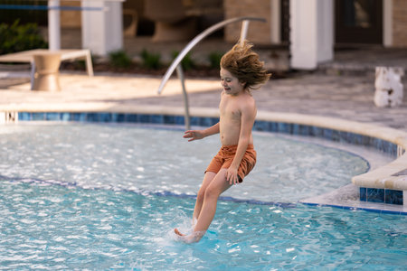 Kid jumping in swim pool. Kid splashing water in pool. Little kid splashing in blue water of swimming pool. Cute child swimming and splashing water with drops in pool.の写真素材