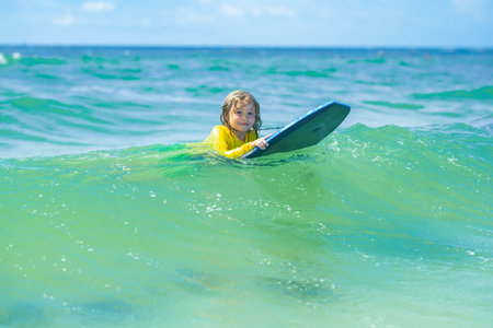 Kid little surfer learn to ride on surfboard on sea waves. Kids outdoor water sport, swimming activity in surf camp. Kid play in summer sea. Kid learning to surf in sea or ocean.の写真素材