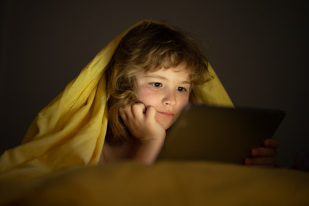 Bed time story. Kid playing game on tablet in bed at night. Kids with social media. Child lying in bed playing a tablet in dark room, light under blacket. Close up of kid watching cartoons on tablet.の写真素材