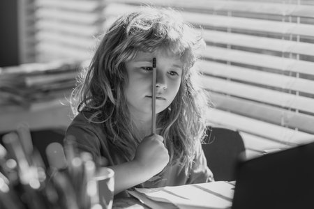 Tired serious schoolboy while doing homework. Portrait school kid siting on table doing homework. Funny child girl doing homework writing and reading at home. Little student at desk in school classroom.の写真素材
