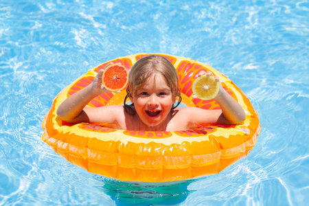 Kid with orange in the pool. Summer fruits. Little kid boy floating in a swimming pool on summer...