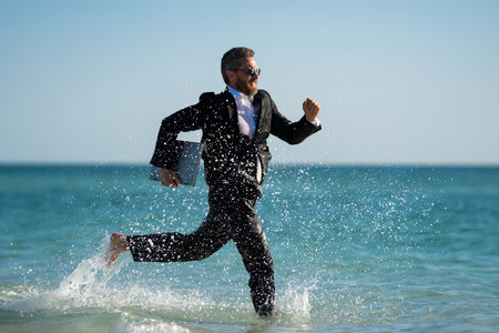 Business man in suit with laptop running in sea water. Travel tourism and business concept. Crazy male office employee with laptop running in sea. Crazy business man on summer vacation.の写真素材
