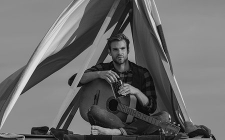 Man playing guitar and sitting near the tent in the camp. Romantic guy camping outdoors and sitting...