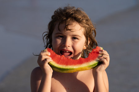 Kid eating watermelon at the beach. Child holding slice of watermelon on beach. Summer fun holiday and travel concept. Child in sea during vacation outdoors. Kid is smiling, enjoying eating fruit.の写真素材