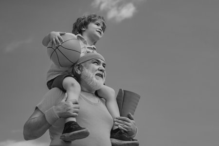 Preparing for morning workout in the park. Happy smiling boy on shoulder grandpa looking at camera. Child sits on the shoulders of his grandfather.の写真素材