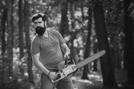 Harvest of timber. Lumberjack concept. Stylish young man posing like lumberjack. Lumberjack worker walking in the forest with chainsaw.の写真素材