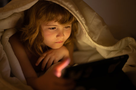 Child gadget addiction and insomnia. Child reads the e-book. Little boy looks at the screen of the tablet at hight. Kid boy playing tablet lying on a bed under blanket. Child social media addiction.の写真素材