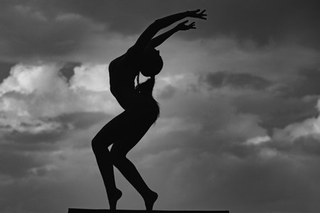 Silhouette of woman stretching. Young girl practicing yoga, doing fit exercise, working out. Dance studio. Cloudy sky background.の写真素材