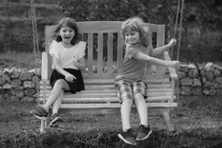 Two children swinging outdoors. Portrait of adorable brother and sister smile and laugh together while sitting on swing outdoors. Happy lifestyle kids.の写真素材