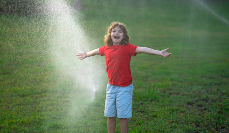 Child play in summer garden. Grass watering. Automatic sprinkler irrigation system in a green park watering lawn. Sprinkler watering. Child gardening concept. Little kid having fun on water spraying.の写真素材