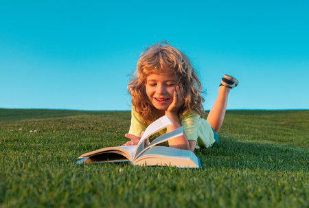 Funny kid reading the book in spring park. Child boy reading book, laying on grass in field on sky background. Portrait of clever kids.の写真素材