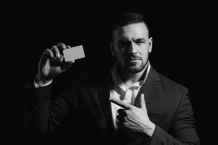 Portrait of a young man dressed in suit pointing finger at a credit card isolated over black background. Businessman with a credit-card.の写真素材