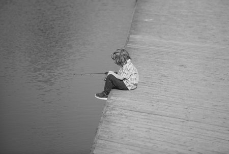 Child boy sitting near the lake and fishing. Kids hobby. Smiling child fishing on the lake. Boy with spinner at river. Boy at jetty with rod, copy space.の写真素材