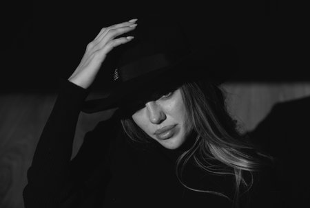 Girl in fashion hat. Young woman posing at studio over black background. Light and shadow. Fashion portrait of beautiful woman with dark light on face. Hidden eyes with wide broad brim hat.の写真素材