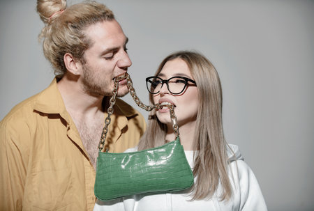 Ladies handbag. Fashion elegant stylish couple in love looking on each other with pleasure. Couple holds fashion handbag in mouth.の写真素材