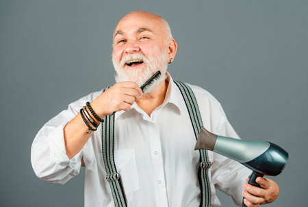 Senior bearded, old man care beard. Portrait of vintage barber. Hair dryer. Retro barbershop, shaving. Hair dresser. Beard care.の写真素材