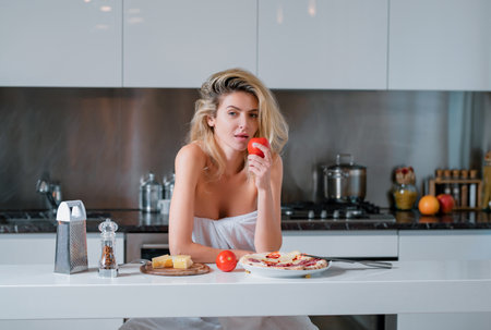 Woman with ingredients for pizza, cheese, sausage and tomato. Young woman eating breakfast. Sexy slim woman at home in morning. Beautiful sensual female housewife in towel with bare shoulders.の写真素材