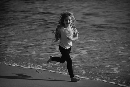 Child running through water close to shore along the sea beach. A boy runs along the sea coast. Rest of children on summer vacation. Little athlete in training. Runner exercising, jogging for kids.の写真素材