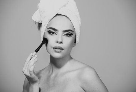 Portrait of a girl with cosmetic brush near face. Woman holds makeup brush. Closeup portrait of a model with bright makeup on a face. Girl with towel on head hold blush blusher, apply powder.の写真素材