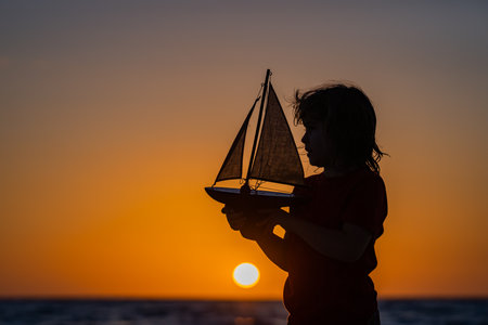 Silhouette of kid playing with toy seailing boat on sunset sea. Little kid boy sailing toy ship on sea water. Summer vacation with kids. Kid dreaming about sailing.の写真素材