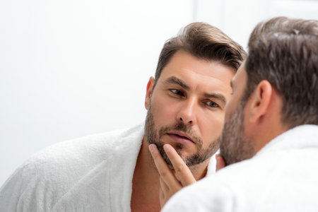 Man applying cosmetic face cream on mirror. Facial treatment. Portrait of man with bare naked shoulders touching skin. Sexy man with skin care product. Male face creme. Perfect skin, morning routine.の写真素材