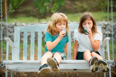 Two kids drink green smoothie outdoors at summer park. Brother and sister happy walking in nature. Siblings boy and girl swinging on swing in garden.の写真素材