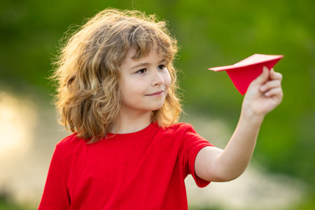 Kid with plane. Cute blonde child dreams fly, playing paper plane outdoor. Growth kids concept. Happy child playing with paper plane in summer garden. Kid throwing paper planes in park. Childhood.の写真素材