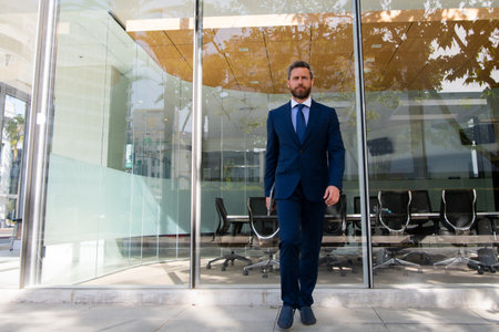 Executive businessman walking. Portrait of ceo near modern office in suit. Happy leader standing in front of company building.の写真素材