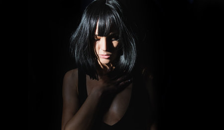 Brunette seductive girl on black background. Young woman posing at studio over black background. Light and shadow. Fashion portrait of beautiful woman with dark light on face.の写真素材
