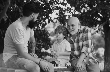 Happy three generations of men have fun and playing Chess in green park. Handsome grandpa and grandson are playing chess while spending time together outdoor.の写真素材