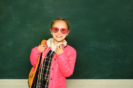 Smiling little schoolkid girl with backpack hold books on blackboard. Childhood lifestyle concept....