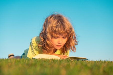 Kid read book in park. Child boy reading book laying on grass on grass and sky background with copy space outdoor.の写真素材