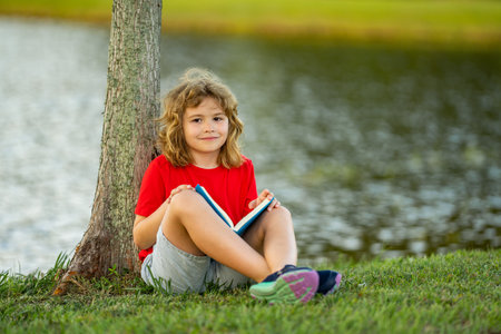 Cute kid reading book on meadow outdoors. School and education. Nature and park. Early learning. Little boy reading a book in summer park. Concept of childrens recreation without gadgets.の写真素材