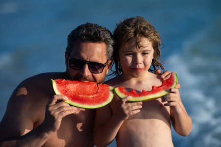 Father and son eating watermelon at the beach. Father and kid son holding slice of watermelon on beach. Summer fun holiday and family travel concept. Father and son in sea during vacation outdoors.の写真素材