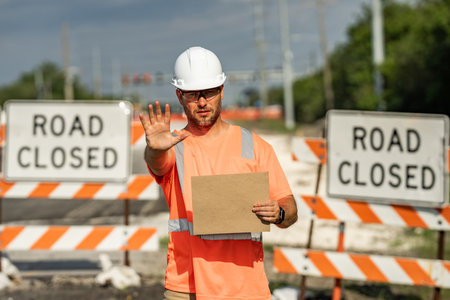 Road closed. Road worker outdoor. Builder in a hard hat working on a construction project at a site....