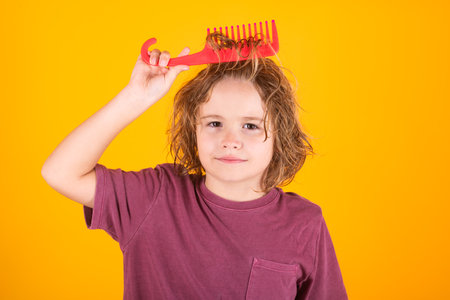 Cute child with curly blonde hair holding comb hairbrush for combing. Cute child with comb. Blonde kid combs unruly hair. Kid boy with tangled long hair.の写真素材