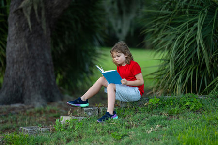 Kid read a book. Early education for kids. Summer vacation with reading book. Outdoor homework. Summer camp with book. Kids reading and education concept. Kid sitting on grass and reading a book.の写真素材
