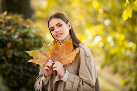 Happy woman holding autumn leafs on face in fall nature. Portrait of young woman with autumn maple...