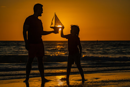 Father and son kid playing with toy sailing boat, toy ship. Travel and adventure concept. Father and son on summer sunset beach. Father and son playing on the beach at sunset. Man family. Fathers day.の写真素材