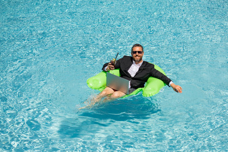 Successful business man floating in suit in pool water. Summer vacations and travel concept. Funny business man drink summer cocktail and using laptop in swimming pool. Summer business concept.の写真素材