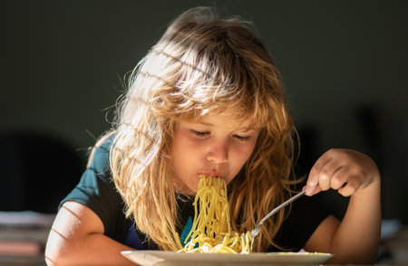 Young kid sitting on the table eating healthy food with funny expression on face. Cute little kid eating spaghetti pasta at home.の写真素材