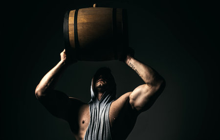 Muscular man wooden barrel on black. Brutal brewer man. Brewery concept. Beer for pub and bar. Oak barrels, keg. Man carries wooden barrel. Barrel with craft beer, whiskey, cognac or wine.の写真素材