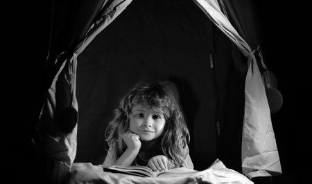 Child boy in kids tent reading book. Serious child reading books, looks serious and concentrated, playing at home, reads story or fairytales.の写真素材
