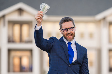 Portrait business man holding cash dollar bills front of house outdoor. Big luck. Dollar cash money concept. Rejoices to win cash. Man hold cash money. Financial luck and success. Manager or realtor.の写真素材