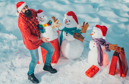 Enjoying nature wintertime. Winter man. Christmas Man and snowman on white snow background. Snowman and funny bearded man the friend is standing in winter hat and scarf with red nose.の写真素材