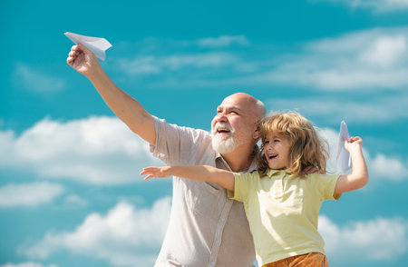 Young grandson and old grandfather with toy paper plane against summer sky background. Child boy with dreams of flying.の写真素材