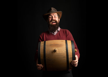 Man hold wooden barrel on black. Oktoberfest. Bearded brewer man. Serious man . Brewery concept. Beer for pub and bar. Oak barrels, keg. Man carries wooden barrel. Barrel with wine.の写真素材