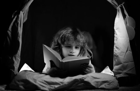 Kid boy in kids tent reading books. Kids face with night light. Concept of education and reading. Kids development of the imagination.の写真素材