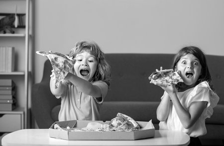 Children eating pizza. Excited kids eating pizza. Two young children bite pizza indoors.の写真素材